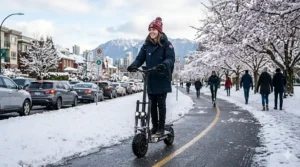 A photorealistic illustration capturing a smiling female commuter wearing a traditional patterned red and white wool tuque (beanie) and a warm navy parka riding a rugged black electric scooter along a cleared, snow-dusted urban multi-use path in Vancouver, BC. Regulatory signs mark the path, contrasting with a long line of fossil-fuel cars stuck and idling in heavy winter traffic on a parallel street.